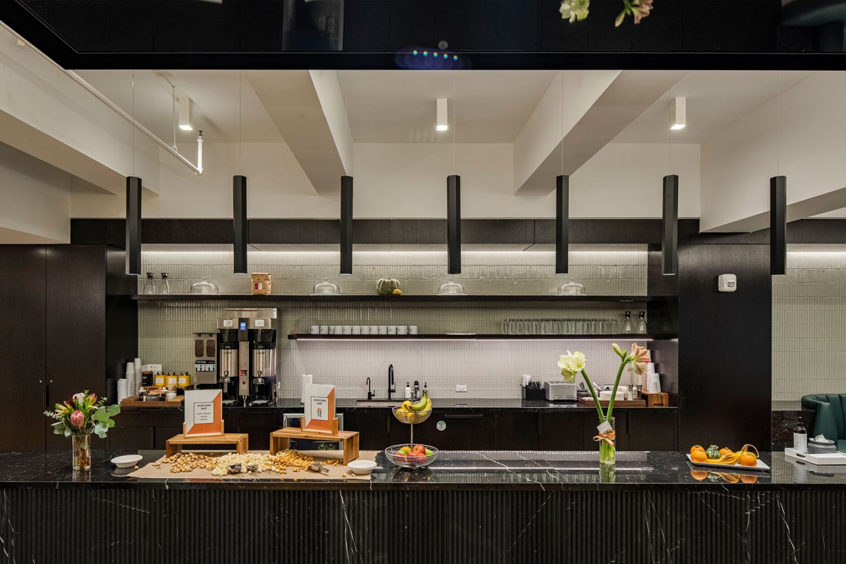 The WorkLife Office Suites by Industrious kitchen area, featuring a long obsidian island table adorned by fruit, snacks, and flowers. In the background are black kitchen cabinets, coffee machines, a sink, and shelves for dishware and cups.