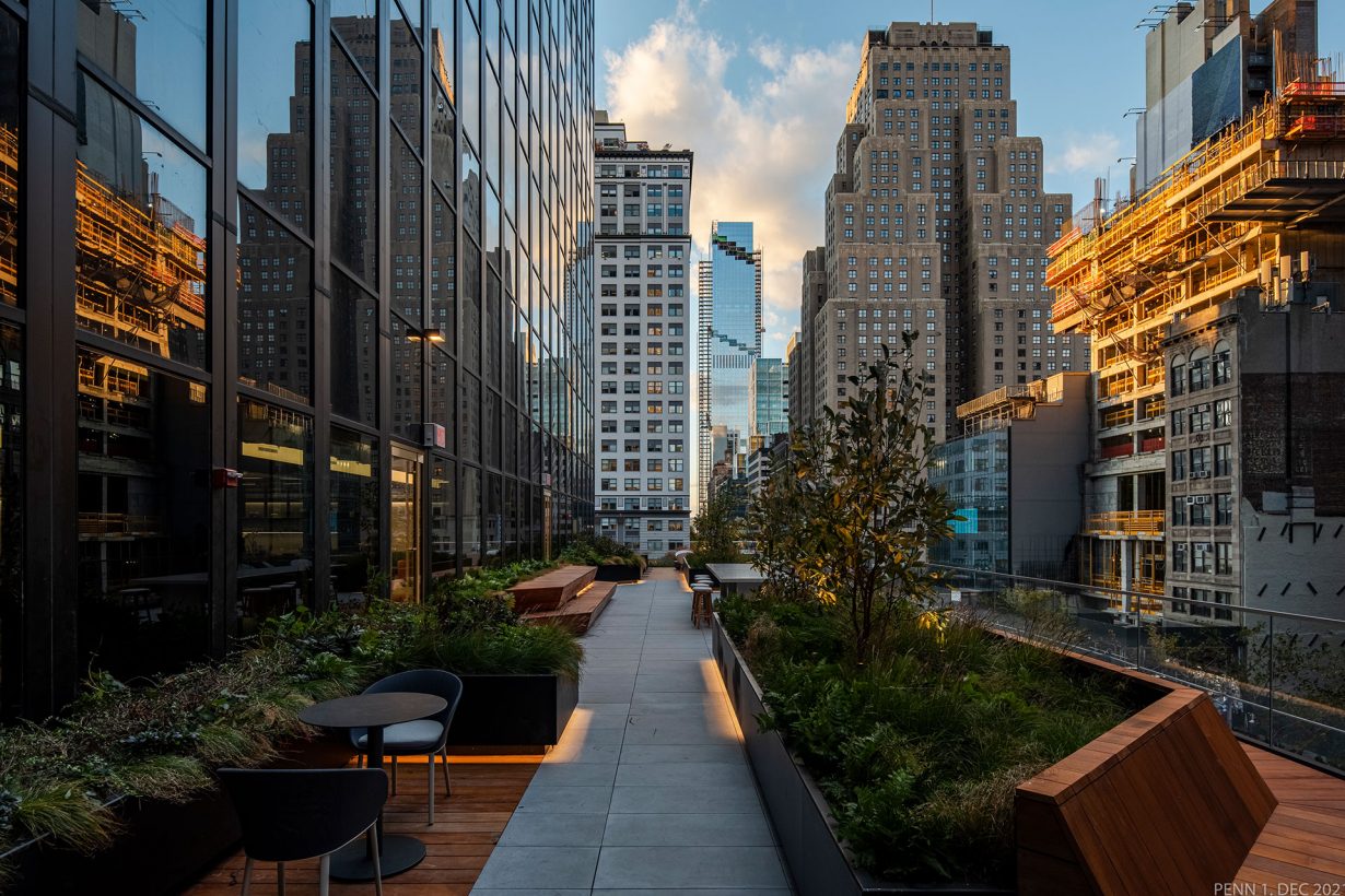 The WorkLife Office Suites balcony terrace, decorated with planters for small trees and greenery, and brown wood benches. The terrace overlooks the city block around PENN1 in New York, with a clear view of sunbeams cast on the surrounding building and a blue sky filled with clouds.