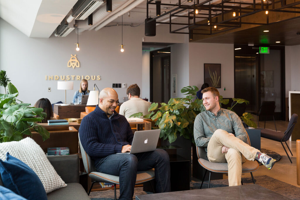 Two men sit in comfortable chairs at a warmly lit collaborative workspace. Behind them, the office manager works on a laptop. On the wall behind her, the company's name reads 
