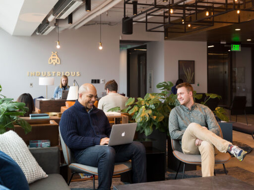 Two men sit in comfortable chairs at a warmly lit collaborative workspace. Behind them, the office manager works on a laptop. On the wall behind her, the company's name reads 