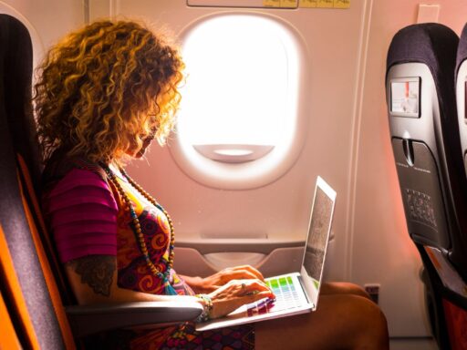 A woman living the lifestyle of a digital nomad is sitting on plane and doing work on a laptop in her lap.